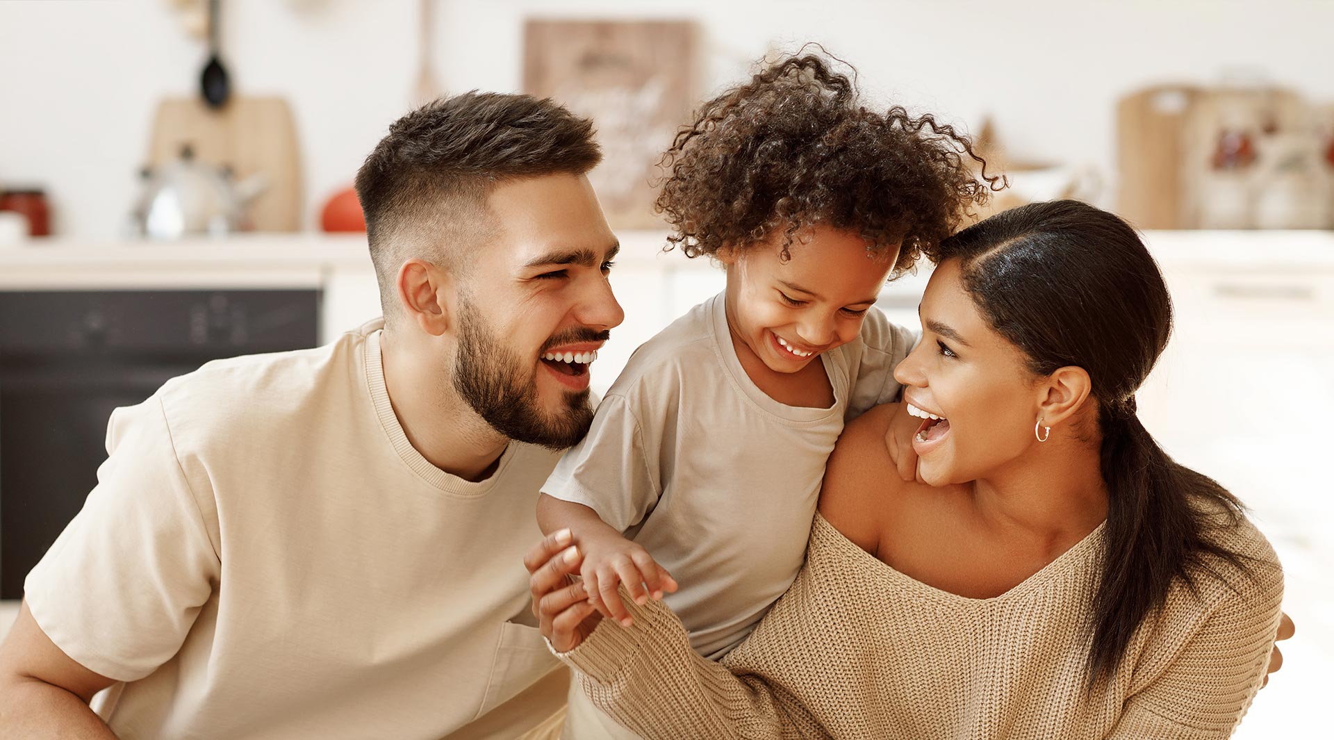 A family of three, including two adults and a child, smiling together in a kitchen setting.