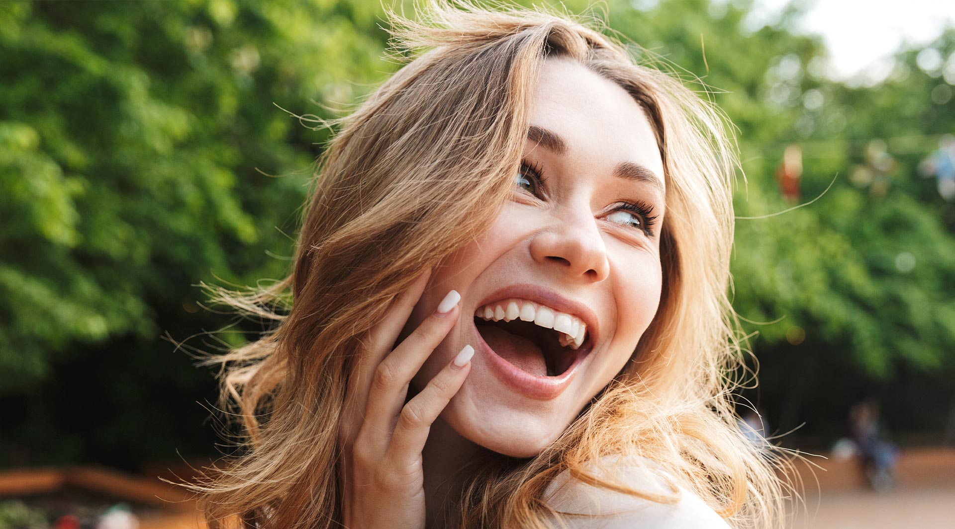 This is a color photograph of a woman with her mouth open, appearing surprised or excited, with her hand on her chest, standing outdoors during daylight.