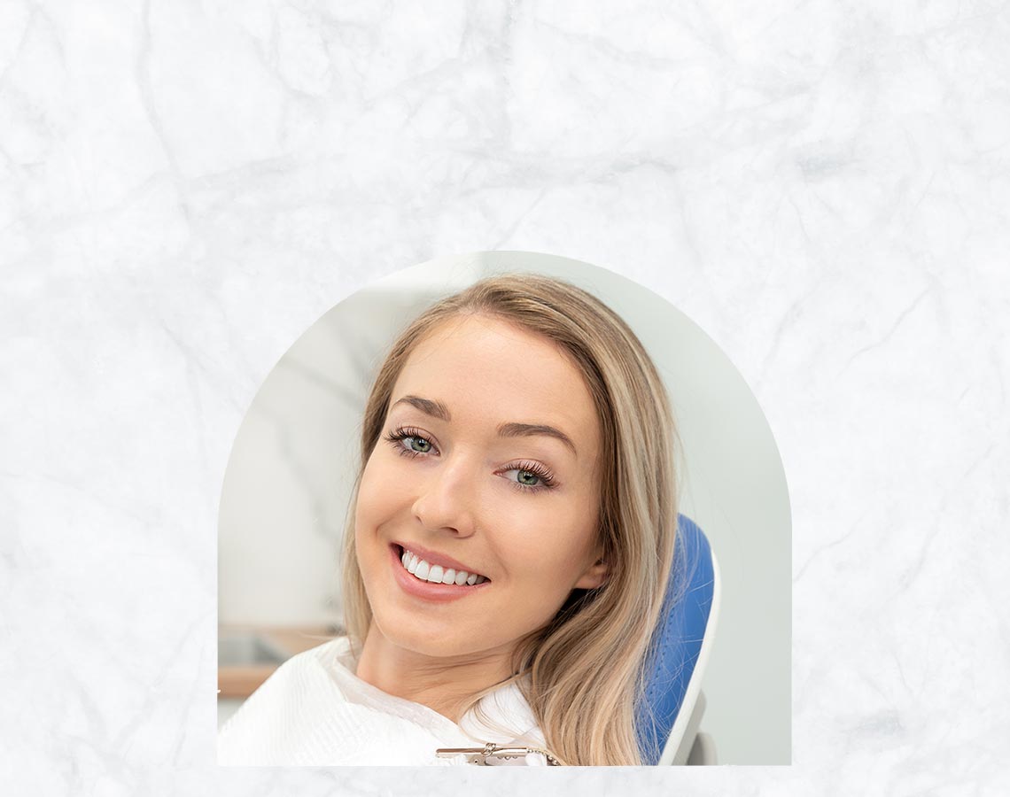 The image shows a woman with blonde hair sitting in a dental chair, smiling at the camera.