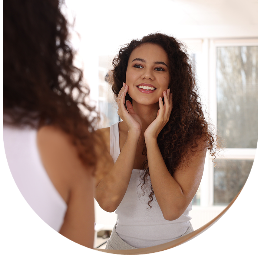 A woman with curly hair smiling at her reflection in a round mirror.