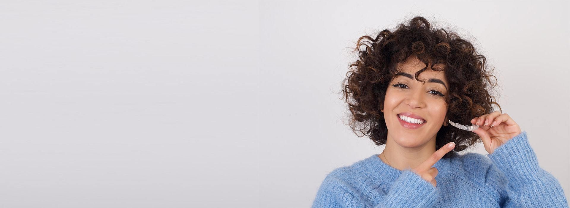 The image shows a person with curly hair smiling at the camera while holding a toothbrush, wearing a blue sweater, set against a white background.
