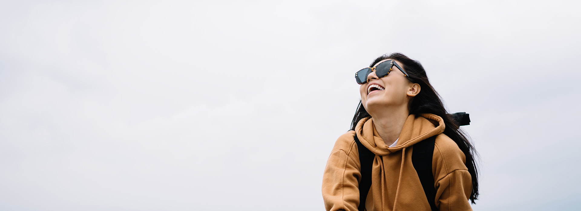 The image shows a person from behind, sitting on a bench with their head turned towards the camera, wearing sunglasses and a backpack, against a backdrop of a clear sky.