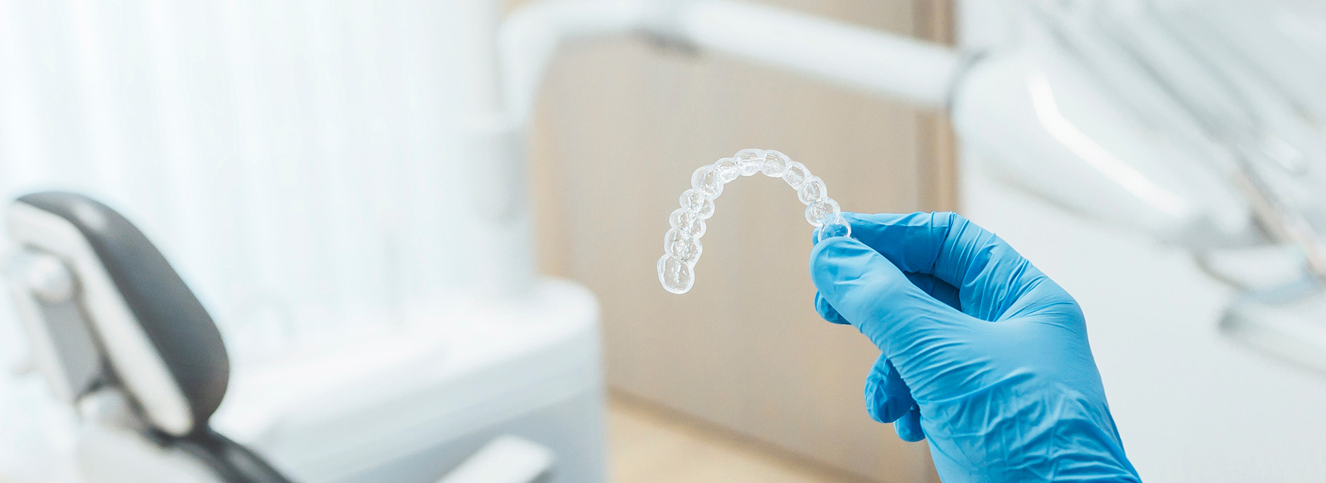 A hand wearing blue gloves holds a transparent plastic bag with a clear white substance inside, set against an out-of-focus background that includes a room with medical equipment.