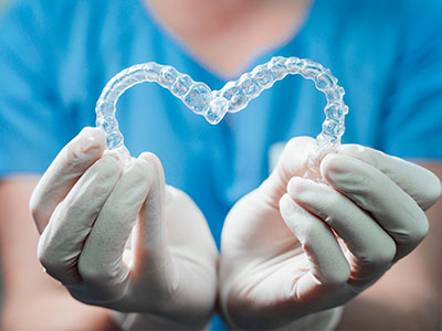 The image shows a person s hands holding a transparent heart-shaped object, which appears to be made from a clear material, possibly plastic or acrylic, set against a blurred background that suggests a medical environment with blue and white colors.