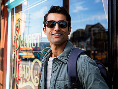 A man wearing sunglasses and a backpack stands smiling outside a storefront window.