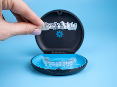 The image shows a person s hand holding a clear tray with a set of transparent aligners, which are dental devices used for teeth straightening, placed inside an open blue case that appears to be a dental appliance box.