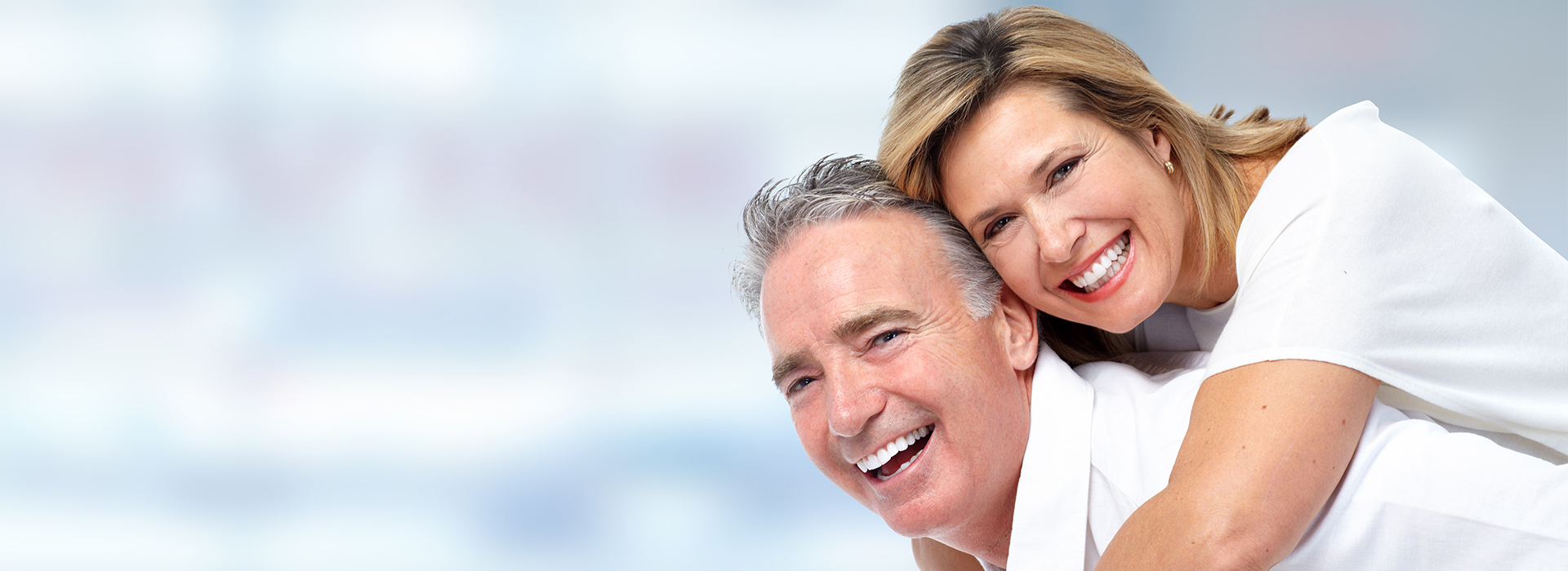 The image depicts a man and woman embracing, with both smiling and looking towards the camera, against a blurred background.