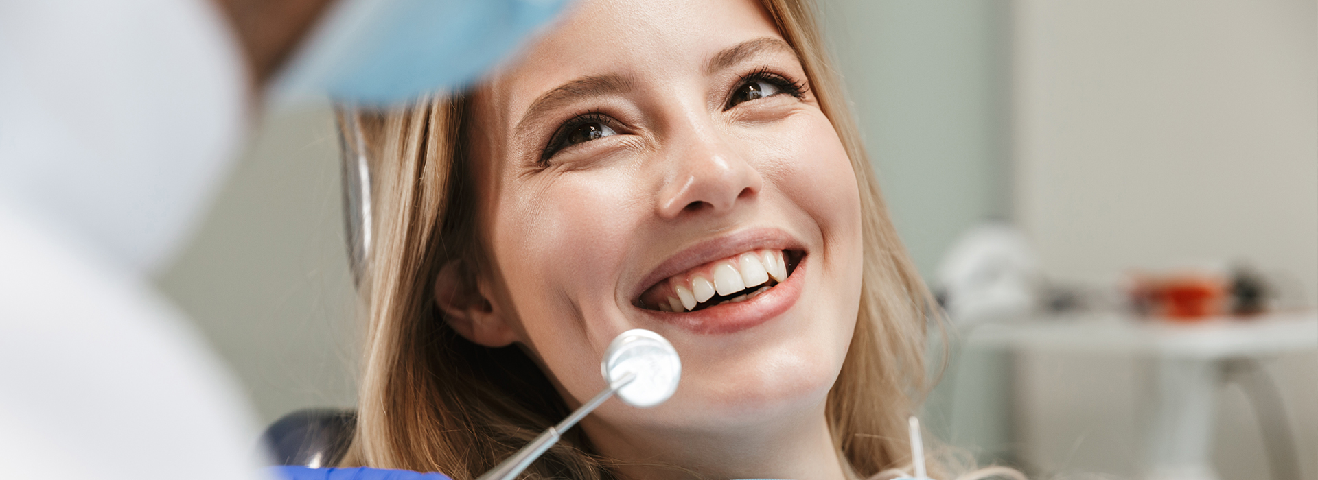 The image shows a young woman smiling at the camera while sitting in a dental chair with a dentist s mirror in front of her, indicating she might be undergoing a dental procedure or consultation.