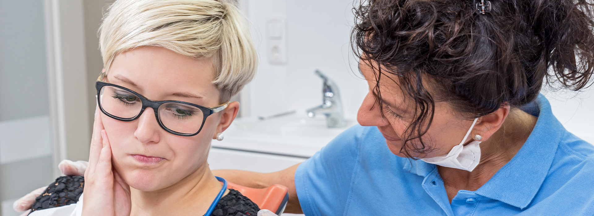 A dentist examines a patient with glasses, both wearing face masks.