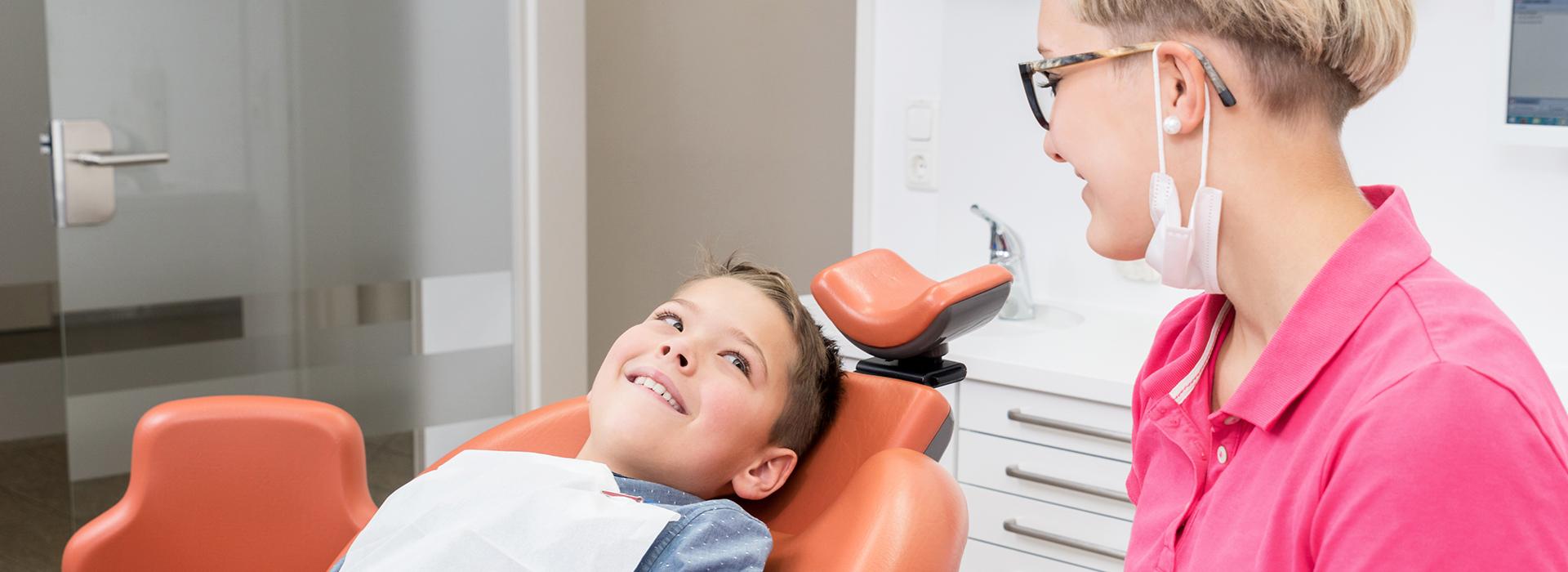 The image shows a dental office setting with two individuals  an adult female patient seated in a dental chair receiving treatment from a dental professional, who appears to be a woman wearing a white coat and a stethoscope around her neck.