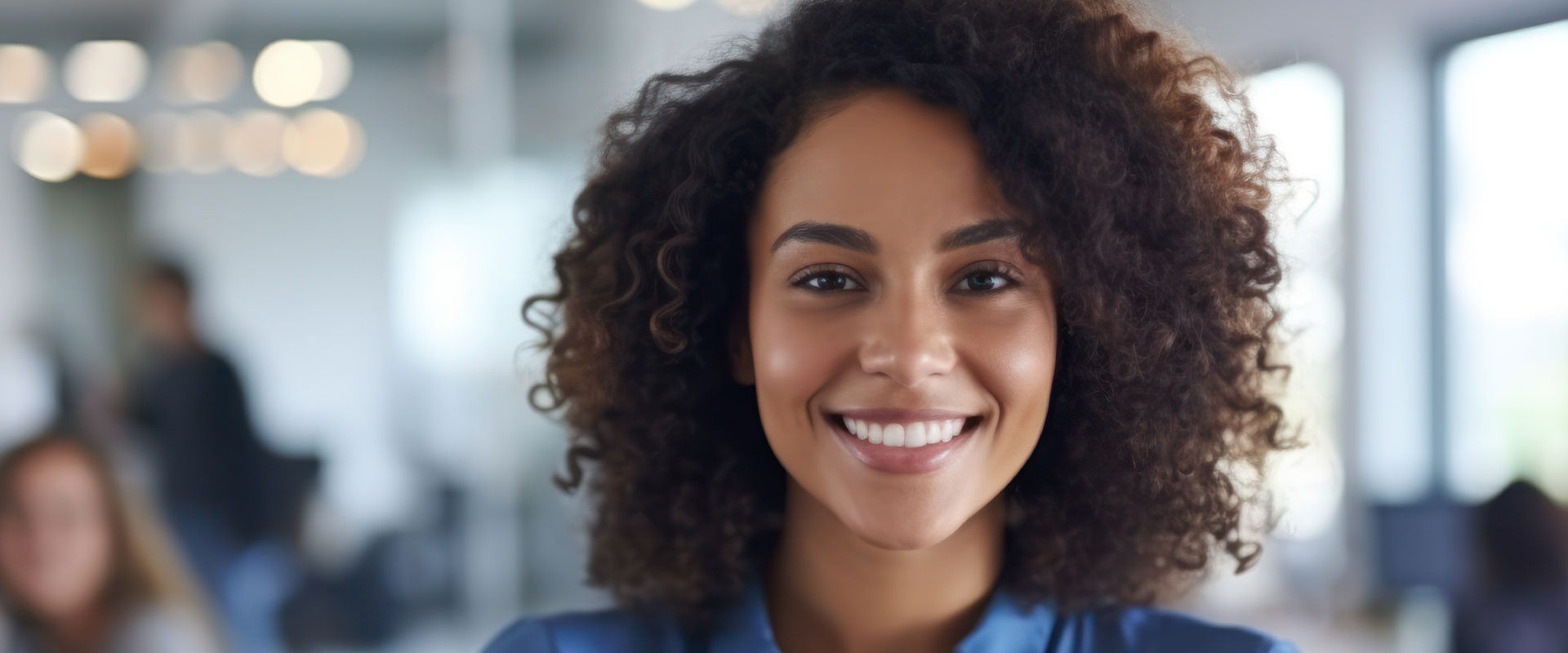 The image features a smiling woman with curly hair, wearing a blue top, standing in an office-like environment.