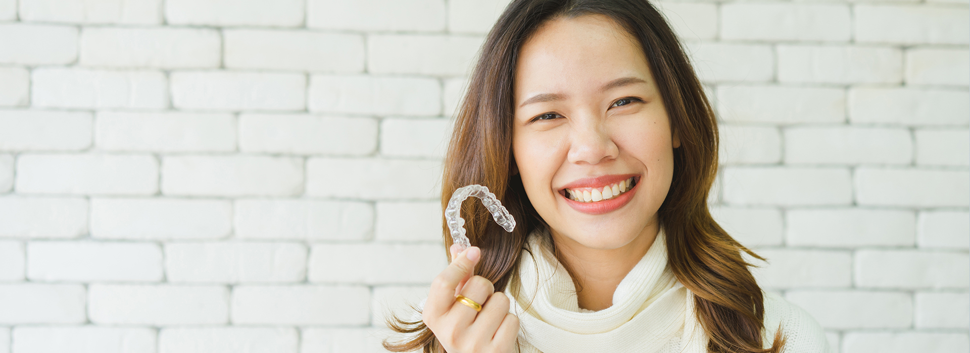 A smiling woman holding up a toothbrush with her right hand against a brick wall background.