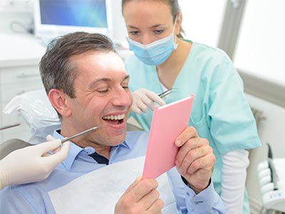 A man sitting in a dental chair with a surprised expression, holding a smartphone, while a dentist and hygienist are observing him attentively.