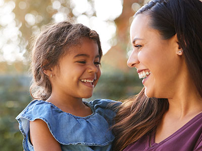 A young girl is smiling at her mother while standing close to her, with both sharing a warm moment outdoors.