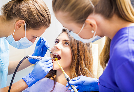 The image shows three individuals wearing medical masks and gloves, performing dental work on a patient who is seated in a chair with their mouth open.