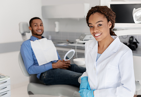 The image shows two individuals in a dental office setting, with one person seated on a dental chair and another standing behind them, both wearing masks, gloves, and protective eyewear  they are smiling and appear to be engaged in a professional interaction.