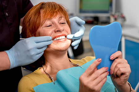 Woman sitting in dental chair with blue mouth guard, holding up mirror with teeth impression.
