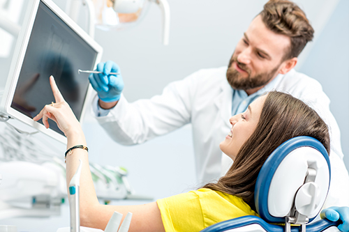 A person sits in a dental chair while a dentist points at something on a computer screen behind them.