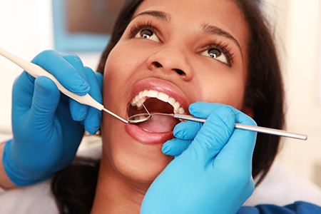 A woman receiving dental treatment with a dental hygienist performing the procedure  she is sitting in a dentist s chair, wearing a blue surgical gown and a hairnet, while holding a mirror to observe her mouth.
