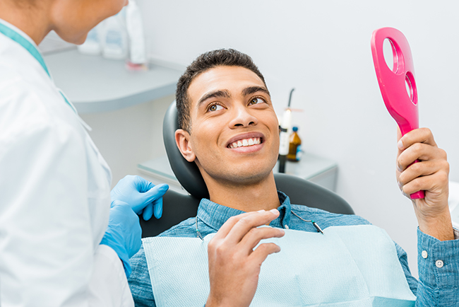 A man sitting in a dental chair with a smile, holding up a pink toothbrush, while a dental professional attends to him.