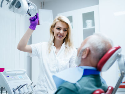 A woman wearing a white lab coat and gloves stands next to an older man with a medical mask over his mouth, who is seated in a dental chair, holding a dental mirror in her hand.
