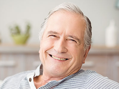 The image shows a smiling elderly man sitting comfortably with his legs crossed, wearing a blue shirt and relaxed in an indoor setting.
