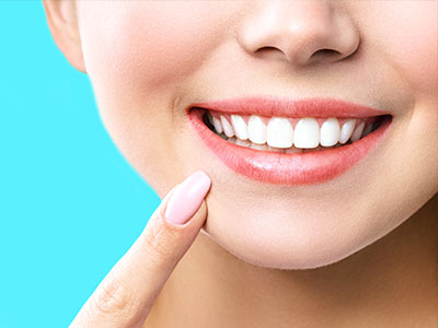 The image shows a close-up of a person with a bright smile, displaying a set of straight, white teeth. The individual appears to be female, with light skin and short hair. She has a small amount of makeup on her lips, and she is holding something up to her face with her right hand. The background is plain and blue, which contrasts with the person s skin tone and highlights their features.